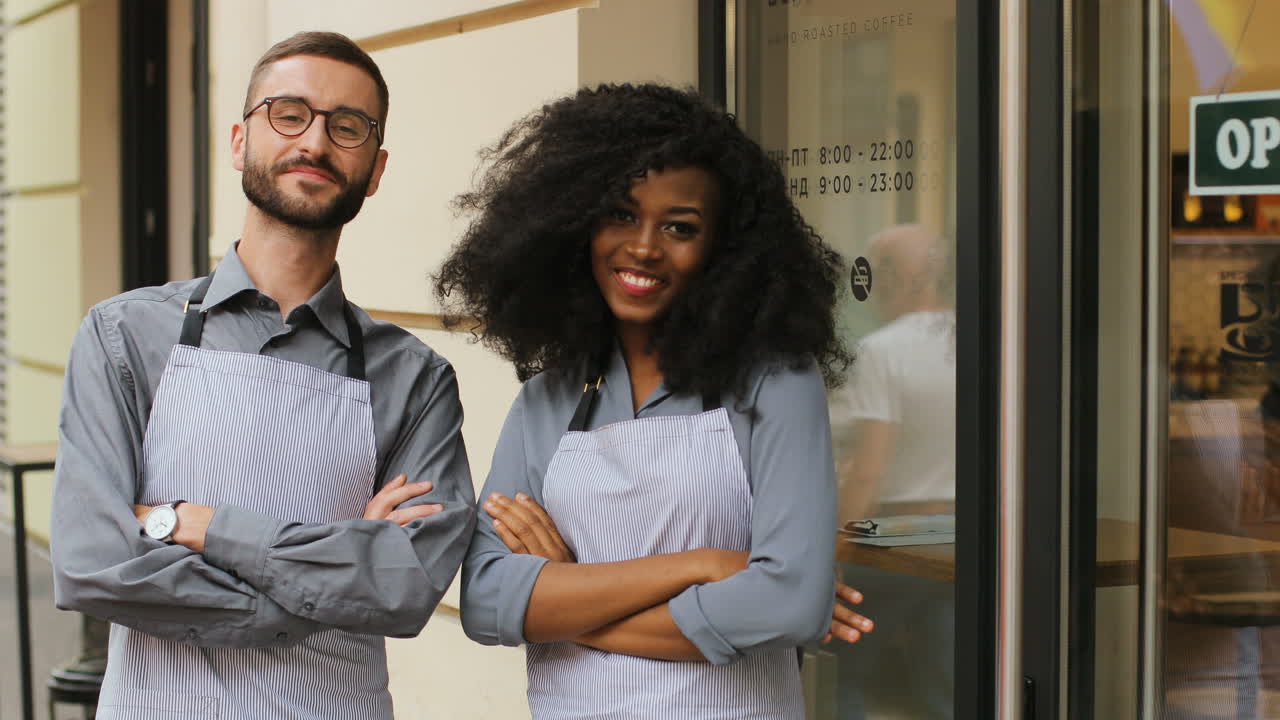 barista caucásico y barista afroamericana de pie en la puerta de un café moderno, sonriendo y mirando la cámara