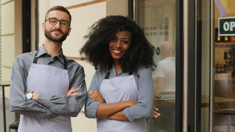 caucasian male barista and african american female barista standing on the door of a modern cafe, smiling and looking at the camera