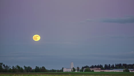 Toma-De-Lapso-De-Tiempo-De-La-Luna-Llena-En-Aumento-En-El-Cielo-Azul-Al-Atardecer-Sobre-Una-Fábrica-Industrial-En-El-área-Forestal-Rural