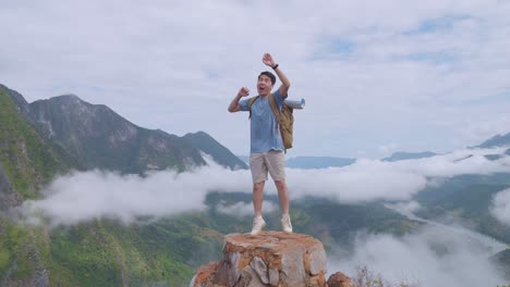 asian hiker male standing on the rock jumping happy and raising his hand celebrating reaching up top of foggy mountain