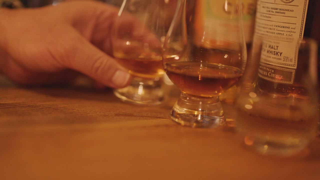 A hand reaches for a whisky glass among several on a wooden table, under warm ambient lighting, with a shallow depth of field and smooth camera movement