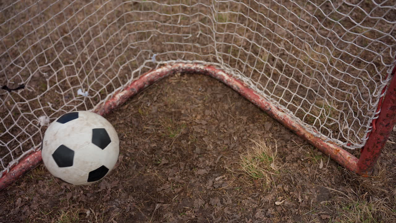 Tomt fodboldmålshjørne med vejrbidt net og rusten stolpe, komprimeret græstørv og plettet græs under dæmpet lys, rolig bybane, der formidler ensomhed og reflekterende efterspil efter kamp, vintage