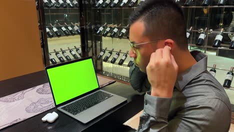 hombre millennial asiático usando auriculares y disfrutando de la música mientras mira una computadora portátil con pantalla verde dentro de su lujosa casa con pantalla de vino