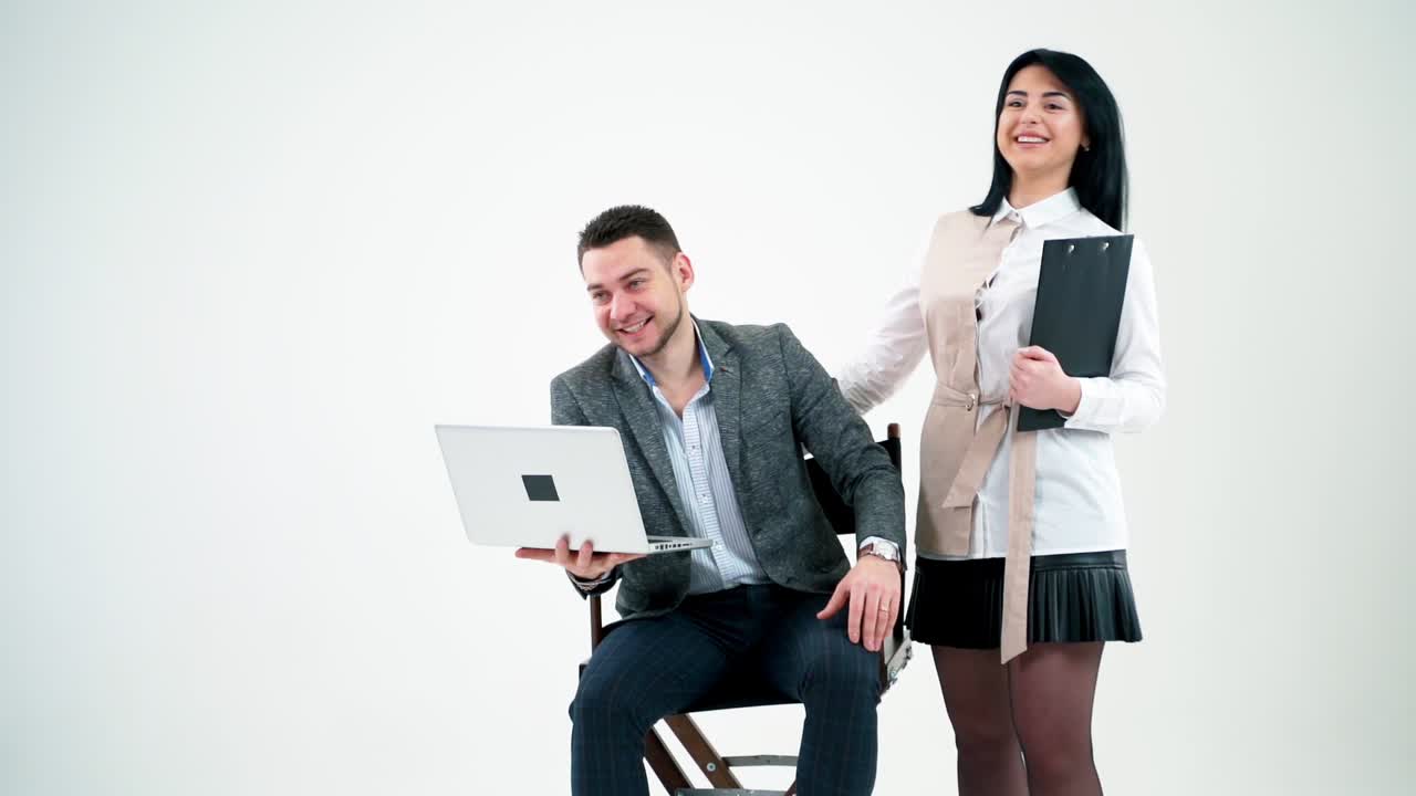 Happy young couple in the studio background. Handsome businessman sitting with a laptop and smiling girl secretary standing nearby.