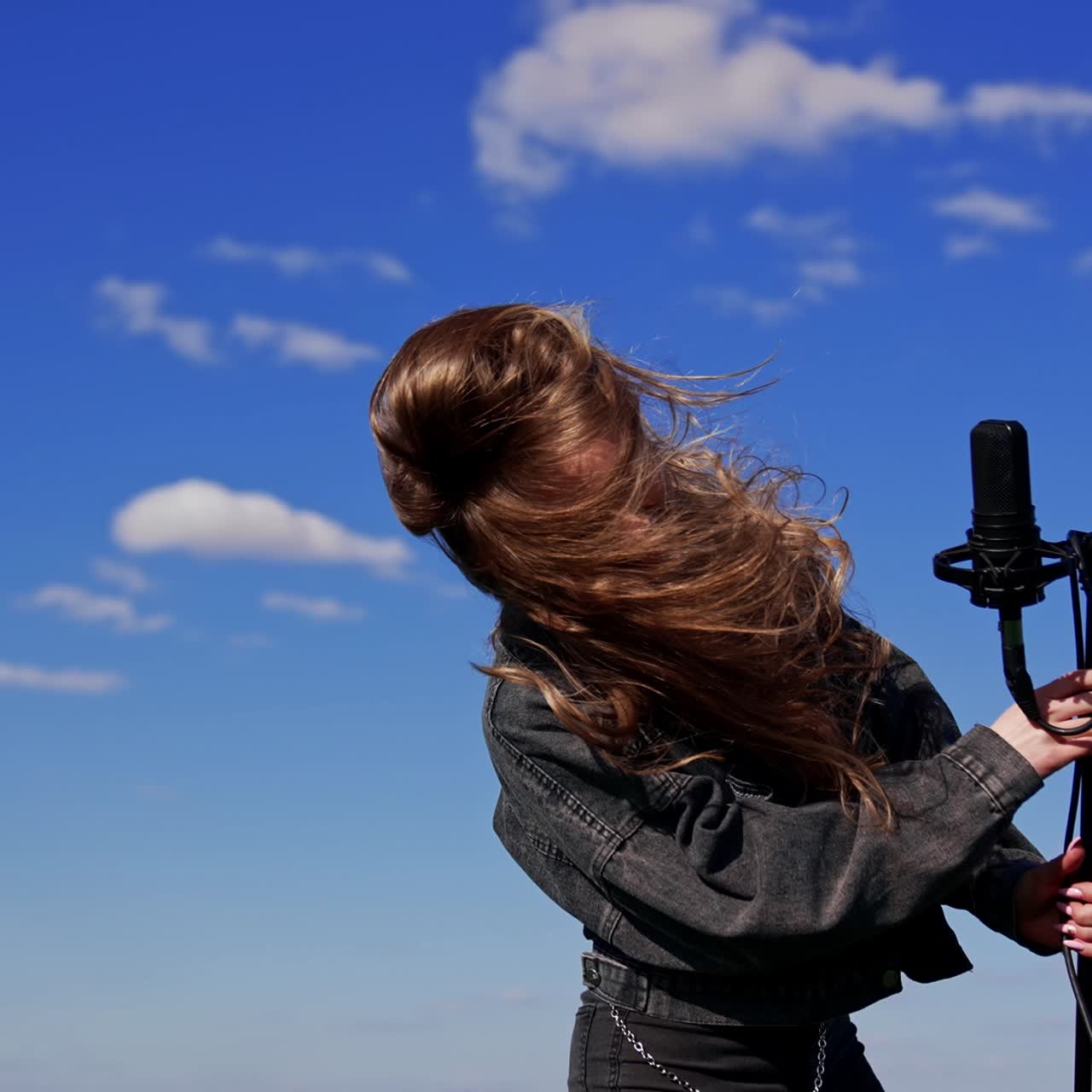 Happy girl singing and dancing in front of microphone outdoors. Female singer moving her head with fluttering hair on blue sky background