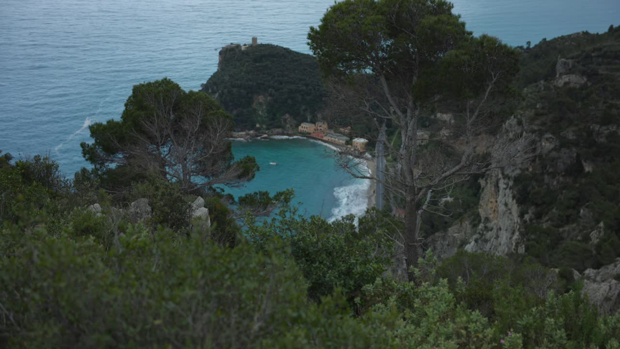Noli, Liguria, Italia: vista panoramica costiera su una baia turchese incorniciata da pini e scogliere, onde su una spiaggia nascosta sotto una fortezza sul promontorio