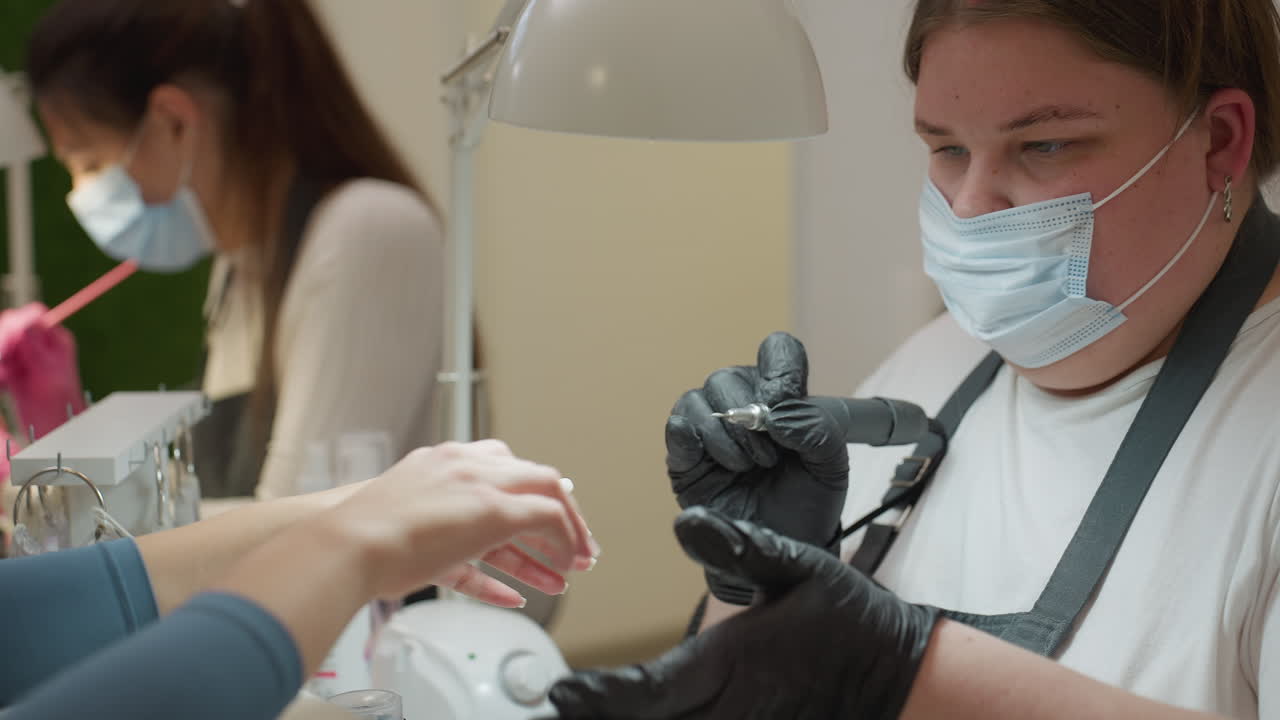Nail technician in black gloves and face mask holding electric nail filer while preparing to file customer nail, with another technician in background attending to different client