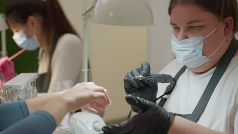 nail technician in black gloves and face mask holding electric nail filer while preparing to file customer nail, with another technician in background attending to different client