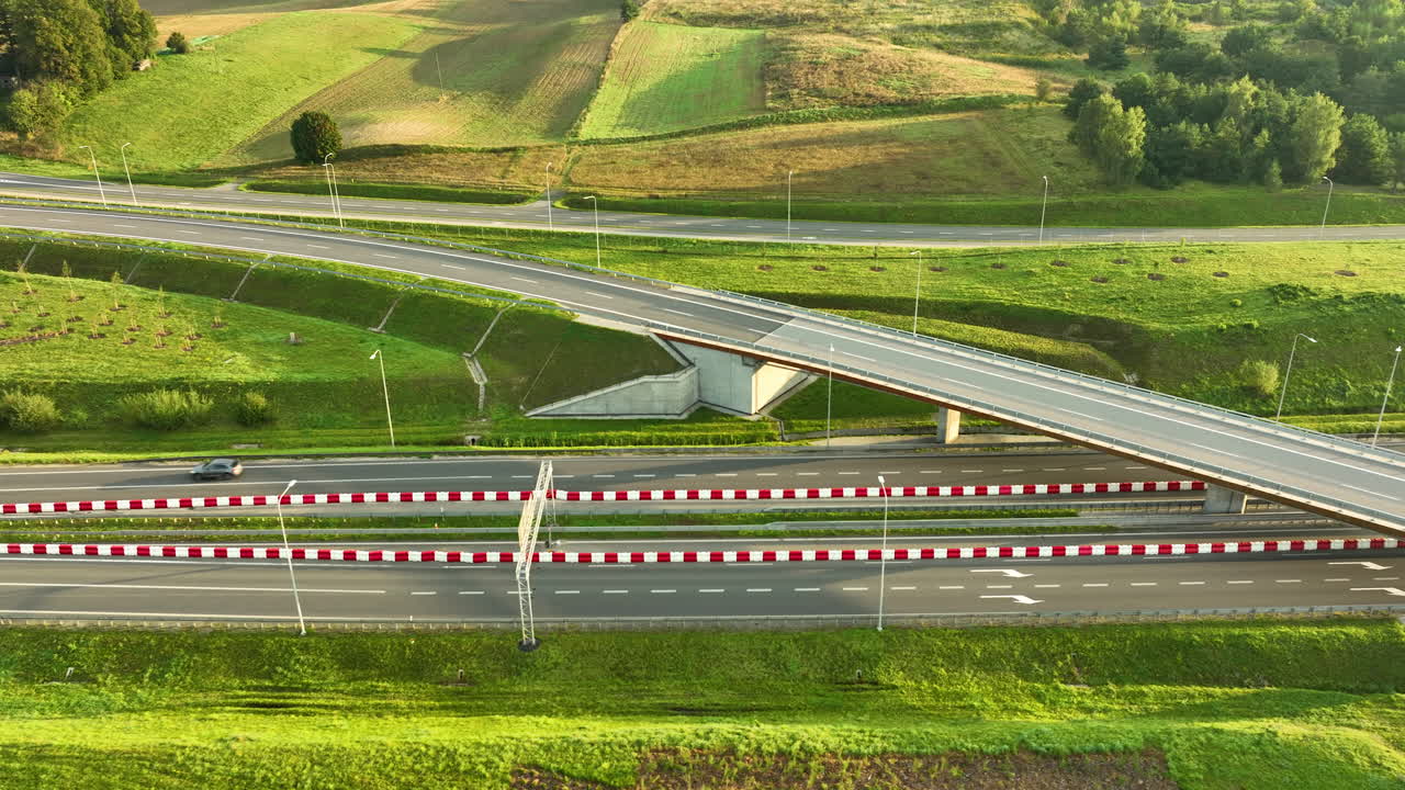 Aerial mid shot focusing on a single white car passing under a concrete highway overpass, flanked by red and white safety barriers and steep green grassy slopes