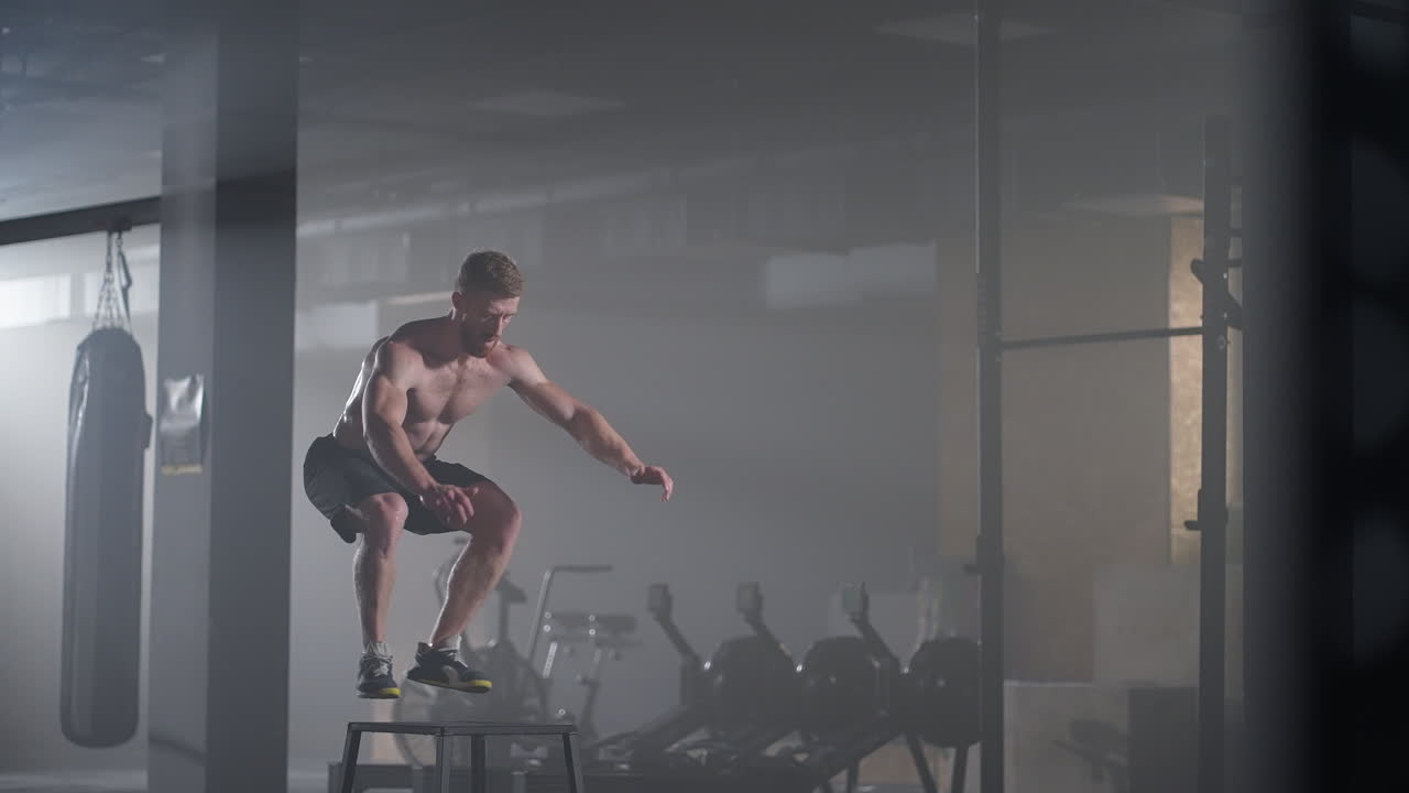Slow motion: Muscular Shirtless Fit Man Energetically Box Jumps in Hardcore Gym doing Part of His Cross Fitness Training Plan. Man is Sweaty from Intense Workout