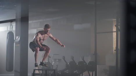 slow motion: muscular shirtless fit man energetically box jumps in hardcore gym doing part of his cross fitness training plan. man is sweaty from intense workout