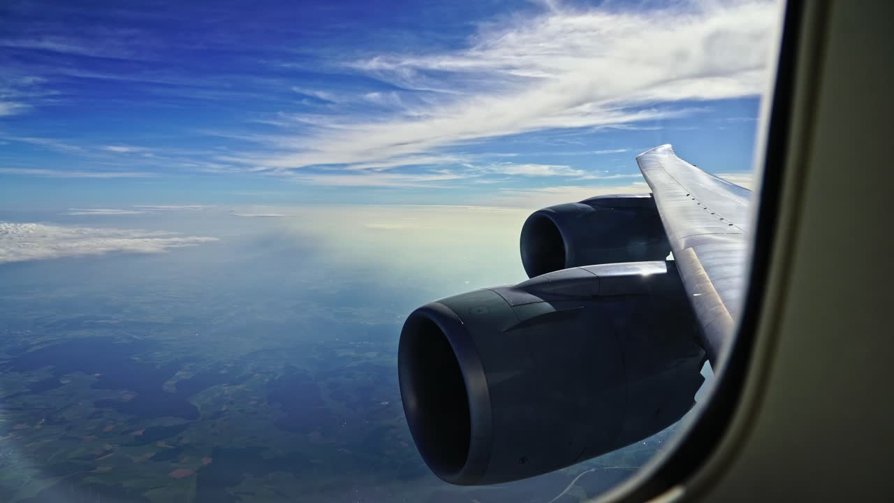 Looking through the window of a passenger airliner with the engines and wing in the foreground against a blue sky with clouds.