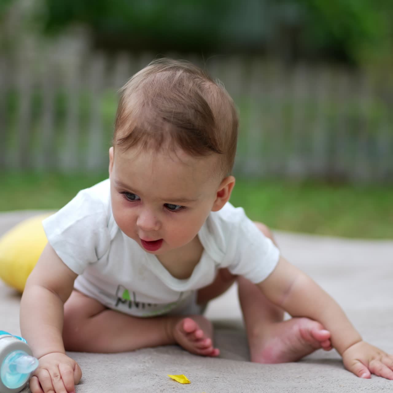 Tiny toddler sitting outdoors playing with his bottle of water. Lovely baby tries to change position to crawl ahead. Blurred backdrop
