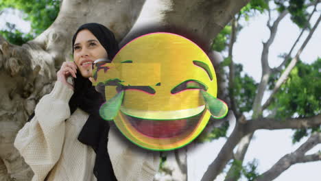 animation of happy emoji over woman in hijab using smartphone