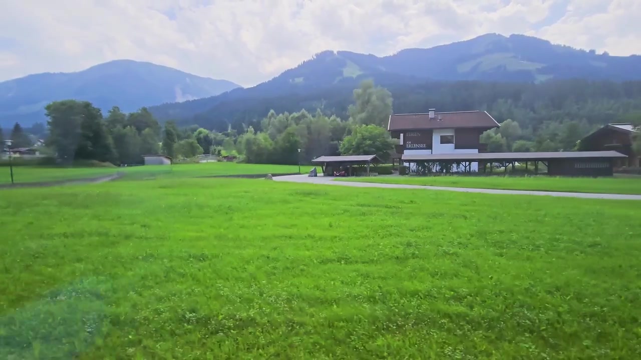 夏のオーストリアアルプスを走行する列車からの眺め。緑の野原、山間の村々、森林に覆われた斜面、そして曇り空が広がり、風光明媚な鉄道旅行や田舎での休暇の映像に最適。