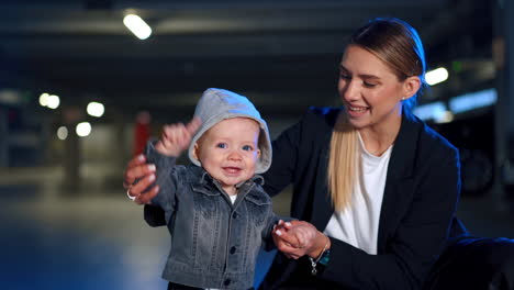 caucasian woman supporting her cute little baby wearing a hoodie and jeans jacket. mom waving kid's hand who's smiling adorably. blurred backdrop.