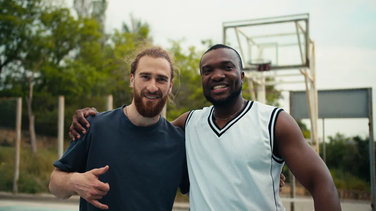 Portrait of two friends, a red-haired man and a black man in a white t-shirt, posing, looking at the camera and smiling on the background of a basketball court