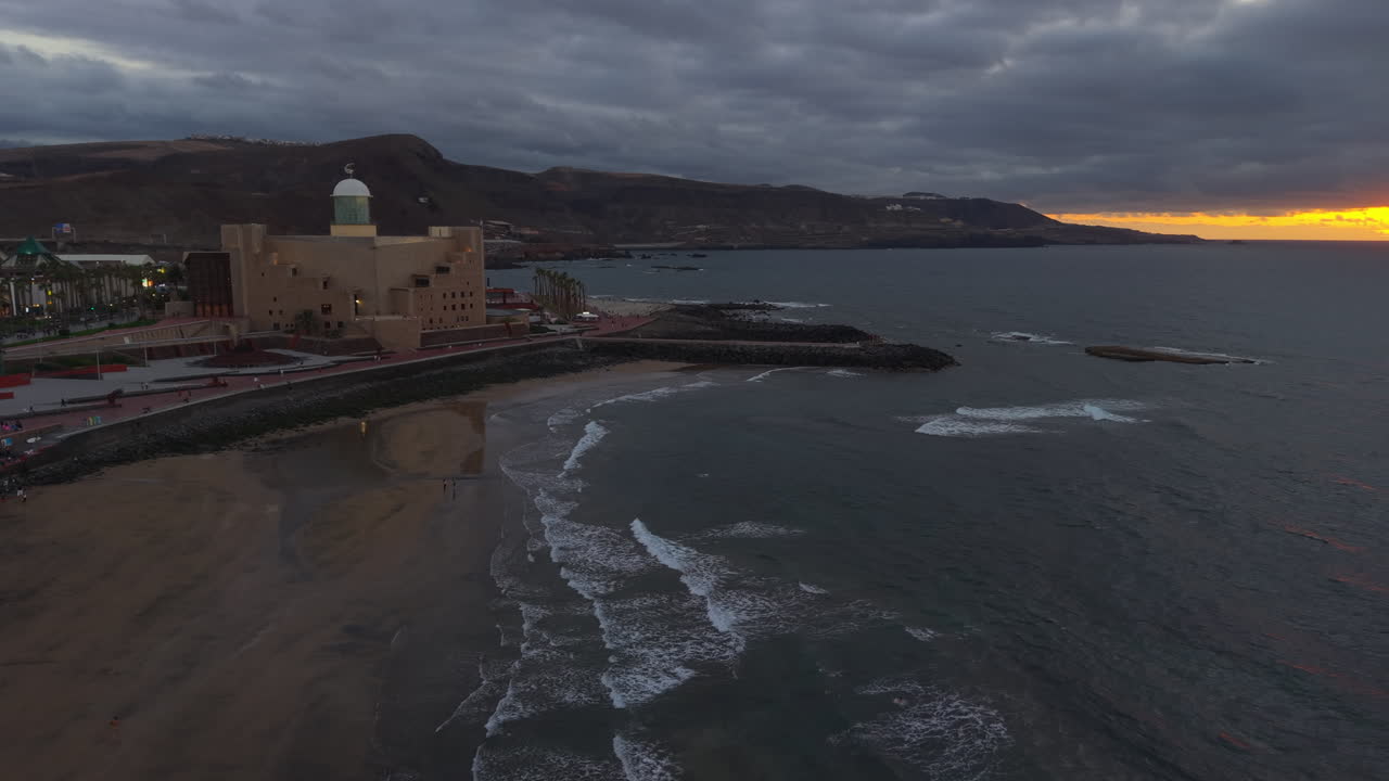 Waves gently washing the shore of Las Canteras beach at sunset, with the Alfredo Kraus Auditorium in the foreground and the city of Las Palmas in the background