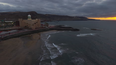 waves gently washing the shore of las canteras beach at sunset, with the alfredo kraus auditorium in the foreground and the city of las palmas in the background