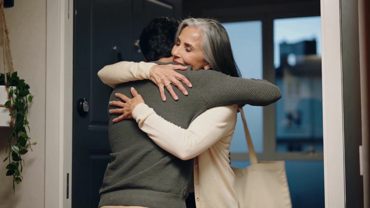 Uomo in arrivo che si gira verso la madre alla porta e l'abbraccia per salutarla, indossando un cardigan e con una borsa a tracolla.