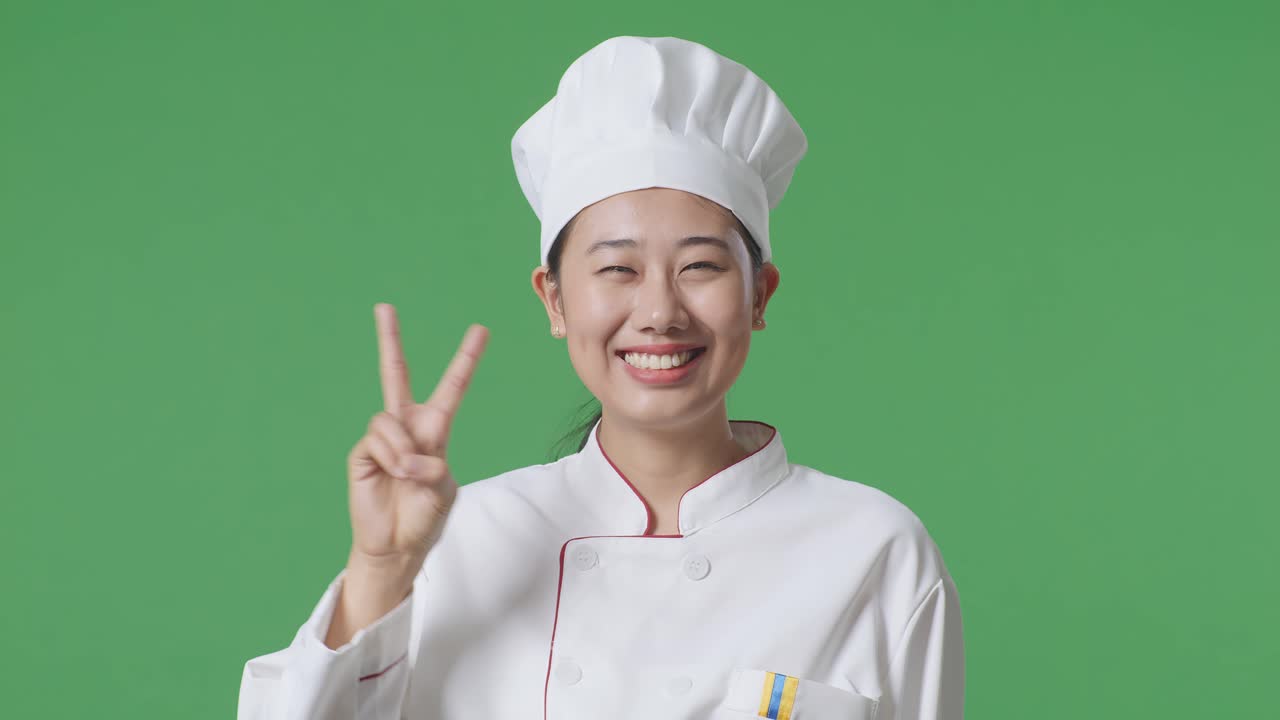 Close Up Of Asian Woman Chef Smiling And Showing Peace Gesture To Camera While Standing In The Green Screen Background Studio