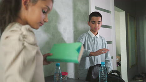 girl boy in casual wear packing notebooks water bottles lunchboxes into backpacks on kitchen counter near fridge before school morning routine siblings organizing healthy meals supplies excited for new day