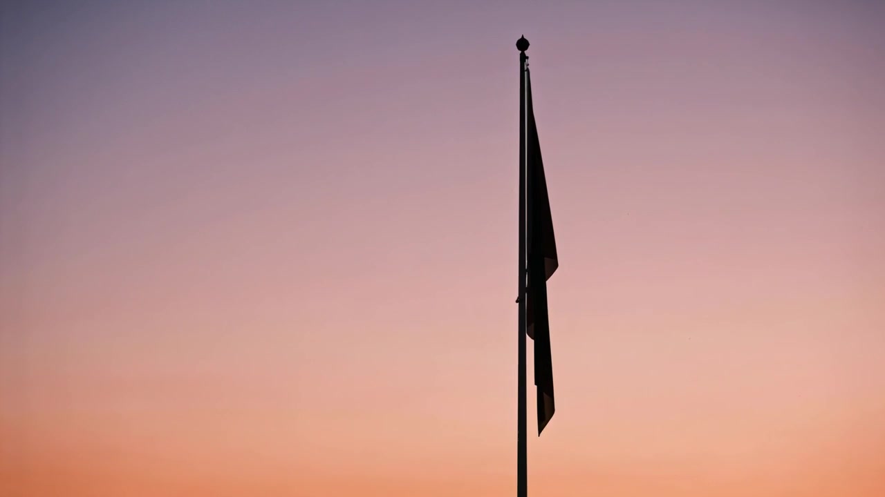 Flatternde dunkle Flagge an einem Metallmast, die auf eine leichte Brise in der Dämmerung reagiert, Flaggenleine vor dem Himmel sichtbar.
