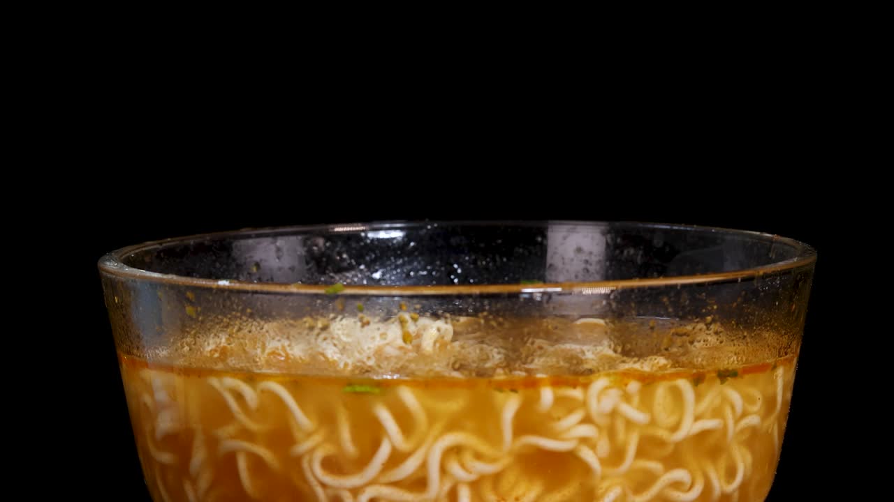 Cooked instant noodles are dropped into a clear glass bowl of steaming broth, with dramatic side lighting and a static camera on a black background