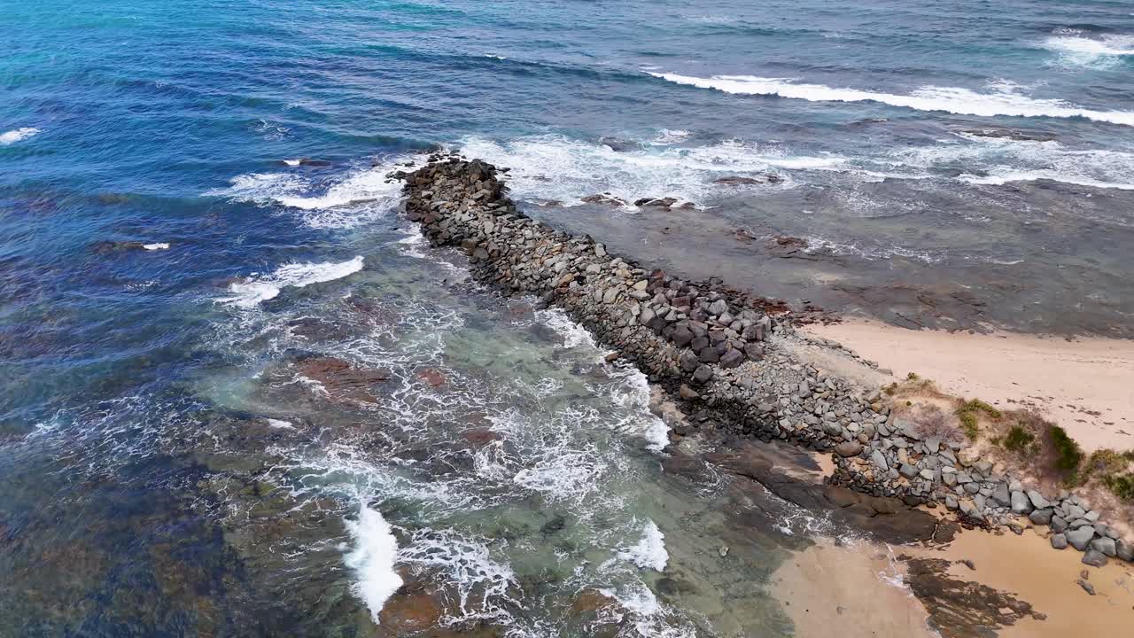 Drone footage captures a rocky wavebreak along Lorne's coastline, showcasing ocean waves and sandy shores under clear skies