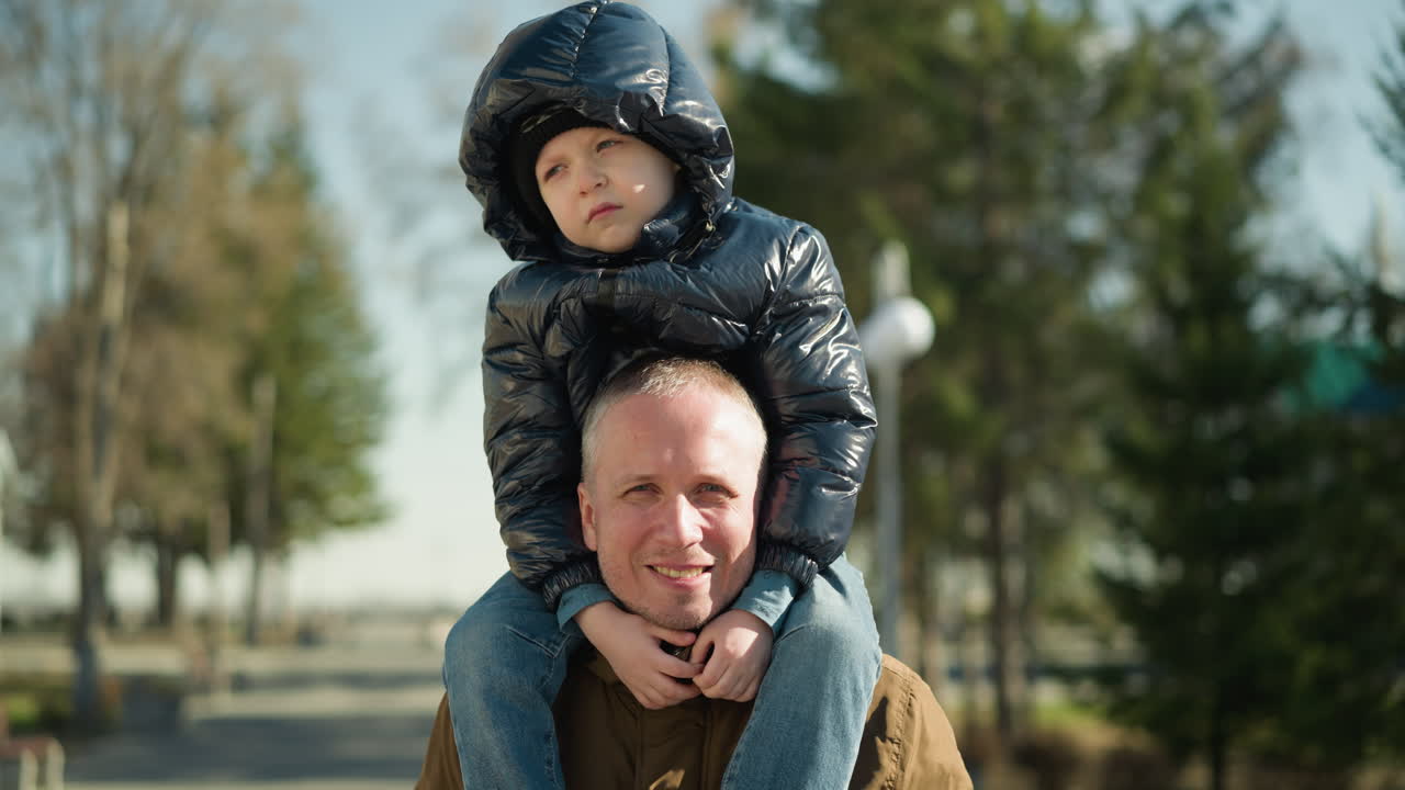 un primer plano de un padre llevando a su hijo en sus hombros sonriendo mientras el niño parece infeliz, con árboles y postes eléctricos detrás de ellos