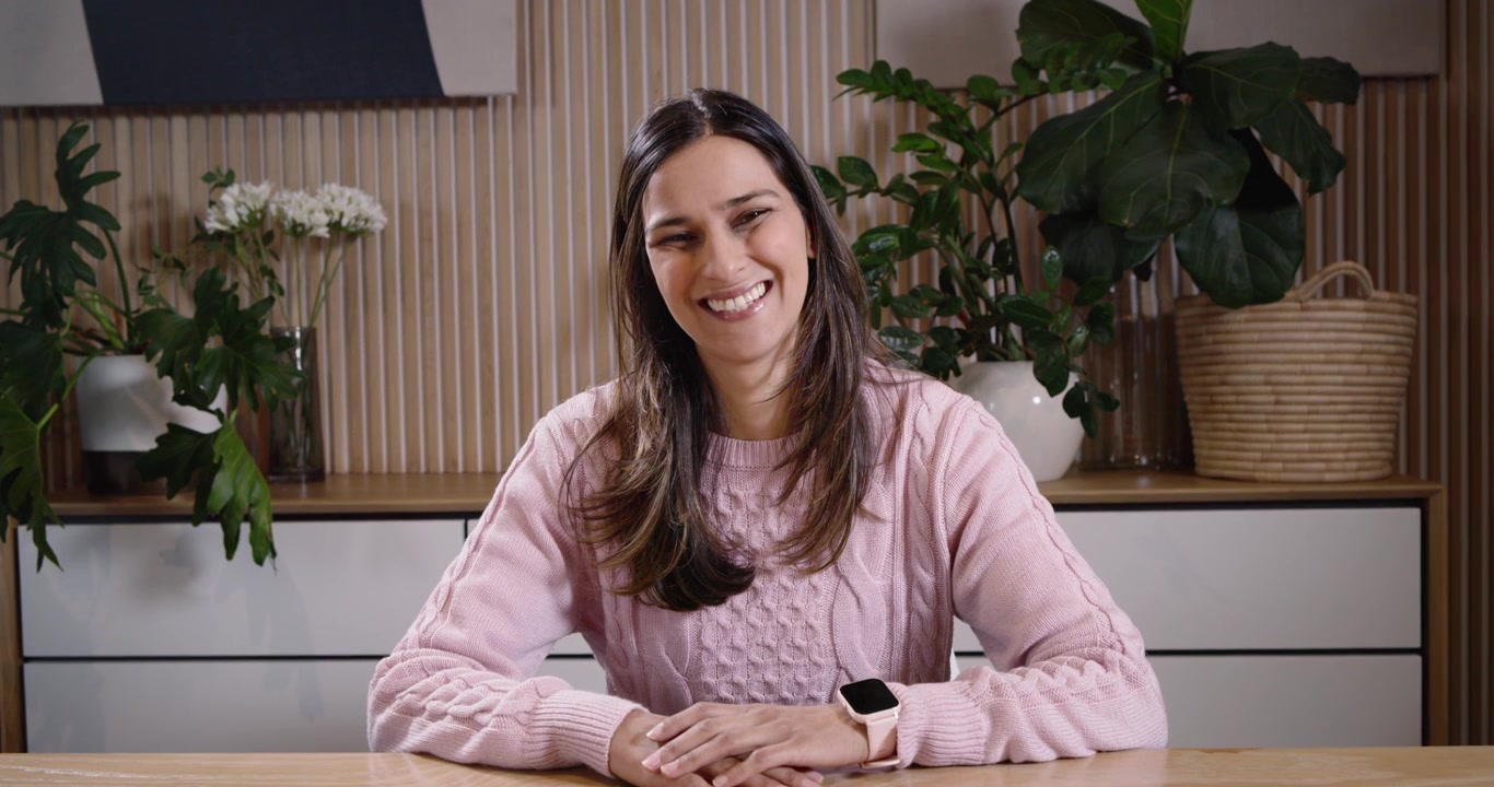 Donna indiana seduta a un tavolo di legno, che posa le mani e poi le mostra alla telecamera, indossando uno smartwatch.