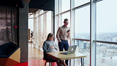pregnant lady working in the office sitting at desk with laptop. woman has a break and touches her belly. male colleague stands beside talking on the phone.