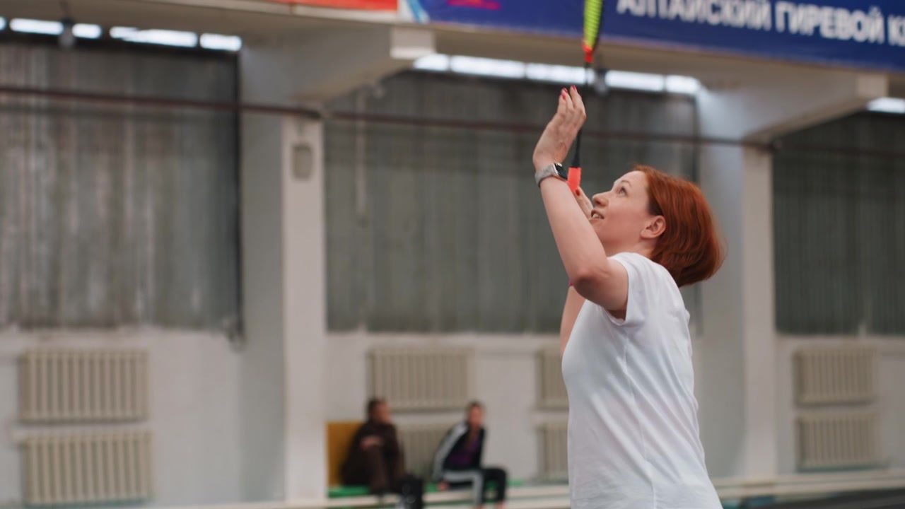 Donna caucasica dai capelli rossi che serve a badminton al coperto, inquadrature dall'alto focalizzate e controllo del polso, che indossa un polsino rosa, esercizio individuale e pratica di precisione, allenatore che osserva a bordo campo, partita determinata