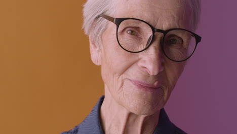 close up view of blonde senior woman with short hair wearing blue shirt and eyeglasses, smiling and looking at camera on orange and purple background