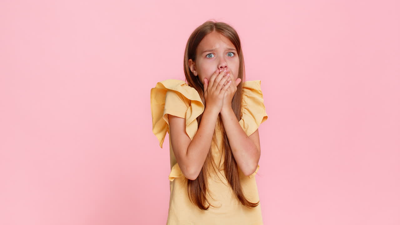 Child girl kid vividly portrays fear emotion covering mouth with hands eyes wide open shocked face