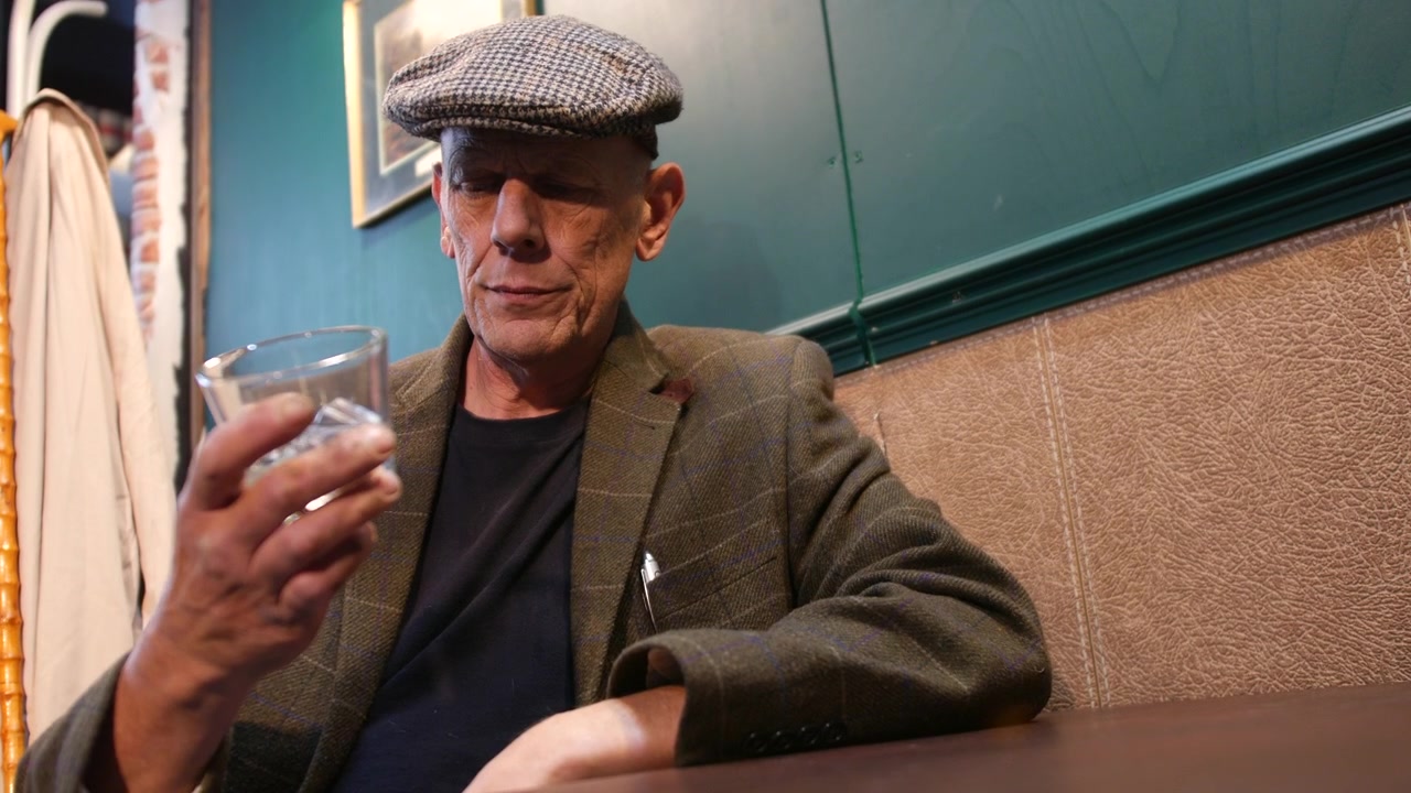 A mature alcoholic man in a flat cap is seated at a table, holding a glass and looking at it thoughtfully and resisting the addiction. His expression suggests deep reflection