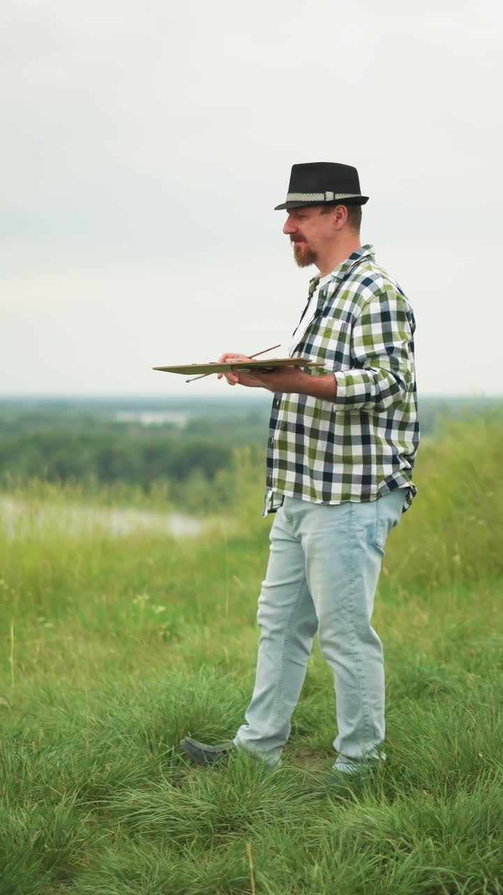 An artist in a checkered shirt, hat, and jeans holds a palette as he walks slowly in a lush grass field beside a serene lake. A black chair is visible in the background