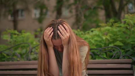 close-up of a woman sitting on a wooden bench, hands covering face, in a reflective, somber posture, her long hair cascades down her shoulders, with lush greenery and trees in the background