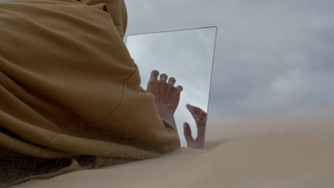 monk in robe finding a mirror in the desert sand dune during a storm and touching his reflection like a mirage