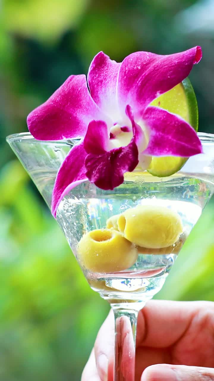 una mano sostiene un martini vibrante con una guarnición de flores, en un telón de fondo exuberante y tropical