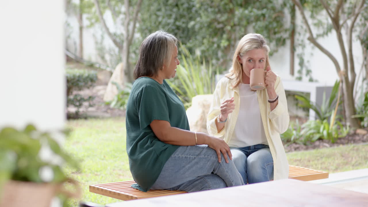 Multiracial senior female friends on bench, drinking coffee, having animated conversation, at home