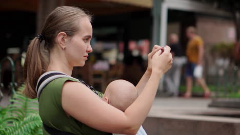 a young mother with her baby in a kangaroo backpack takes photos on a mobile phone while traveling. she walks and glances at the phone screen intermittently