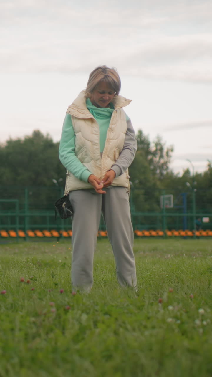 잔디가 있는 공원에서 하얀색 옷을 입은 여성이 보상 가방을 챙기고 있어요. 민트색 후드티와 크림색 조끼를 입고 캔버스 가방에서 간식을 준비하는 동안 작은 강아지가 기다리고 있어요. 흐린 하늘, 잔잔한 아침, 평범한 노년의 일상이에요.