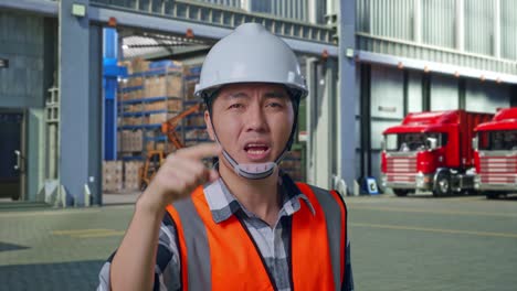 close up of angry asian male engineer with safety helmet shouting at someone while standing , outside of logistics distributions warehouse