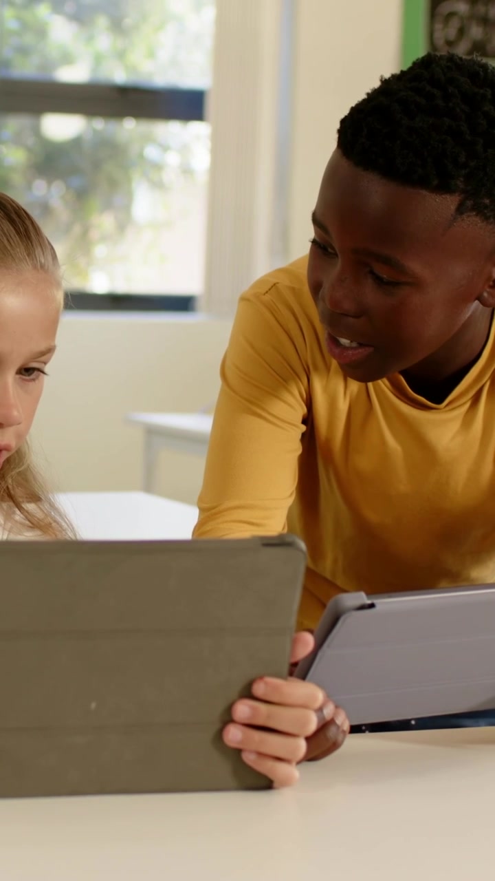 Vertical video: Entering frame, boy in mustard top leaning at school desk aligning tablets to share