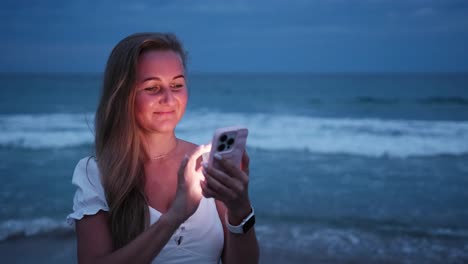 a girl joyfully uses her smartphone by the serene sea during twilight, softly illuminated by the screen