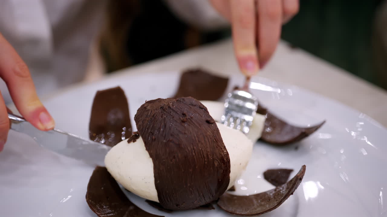 Close-up de uma sobremesa em forma de coco com uma casca de chocolate sendo cortada para revelar um interior cremoso.