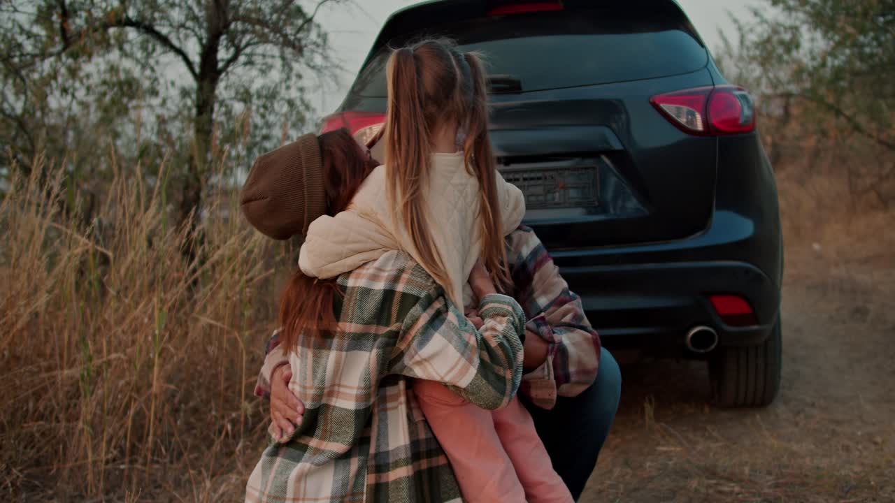 una pequeña chica rubia abraza a sus padres cerca del coche en el que llegaron para un picnic fuera de la ciudad por la noche en el verano. una familia feliz abrazándose durante su picnic afuera de la ciudad en una noche de verano