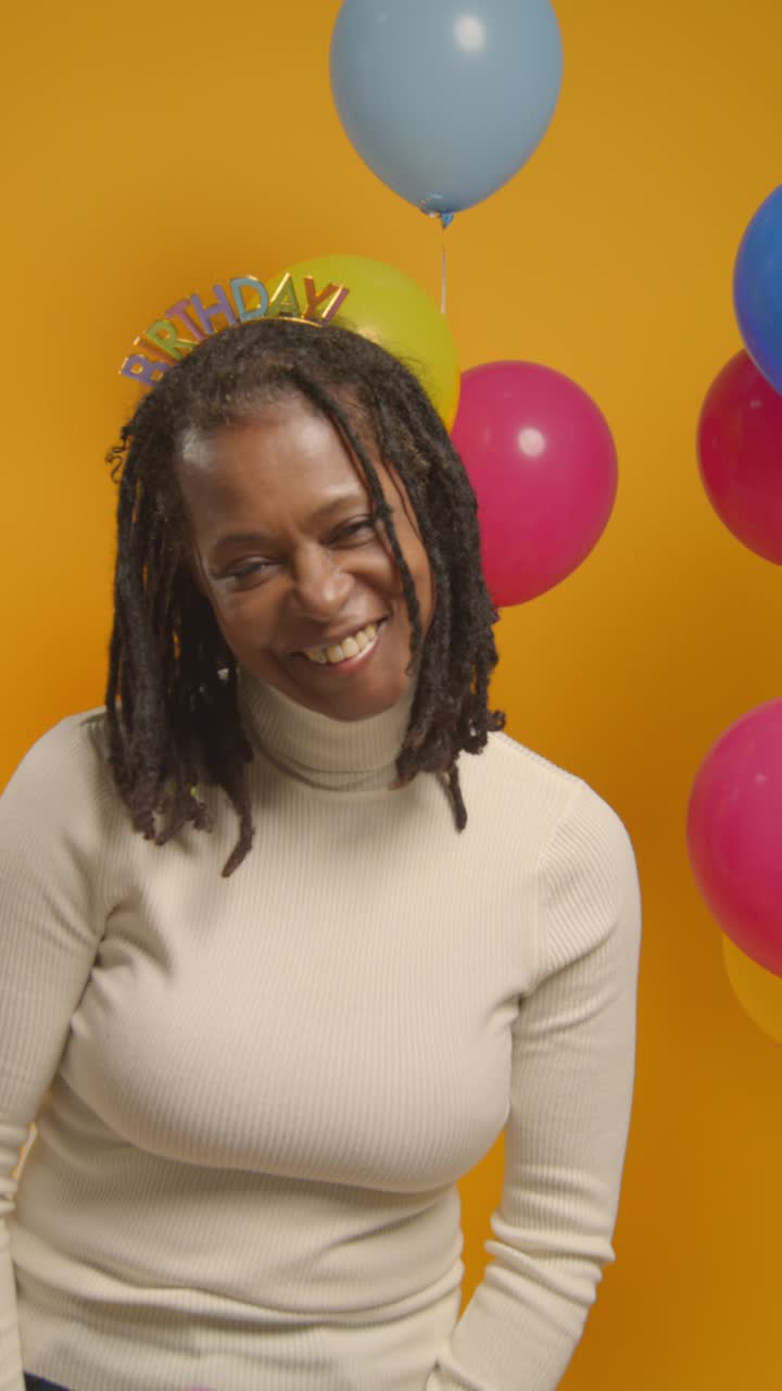 Vidéo verticale : Portrait en studio d'une femme portant un serre-tête d'anniversaire, célébrant en soufflant des confettis en papier vers la caméra