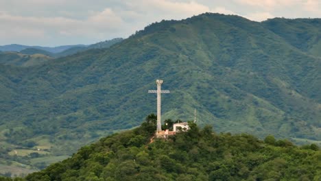 Aerial-view-of-a-grand-cross-monument-amidst-green-mountainscape,-valley,-and-cloudy-sky